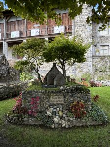 A quaint garden scene featuring a small, decorative stone structure resembling a house, surrounded by carefully arranged stones and colorful plants. In the foreground, red and green foliage adds vibrancy, while a couple of small trees provide shade.