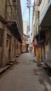 A narrow paved alley lined with old multi-story buildings, traditional balconies, overhead utility wires, and shuttered storefronts, with a scooter parked along the quiet street under a partly cloudy sky.