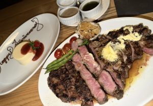 Sliced steak with butter on a white plate with green beans, tomatoes, and garlic. Next to it are small sauce cups and a dessert plate with "Happy Anniversary" written in chocolate.