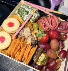 A square snack box filled with an assortment of sweet and savory treats. It includes folded salami, a herb-coated cheese ball, grapes, strawberries, various crackers, jam-filled cookies, wafer bites, pink Pocky sticks, and a gold-wrapped chocolate truffle.