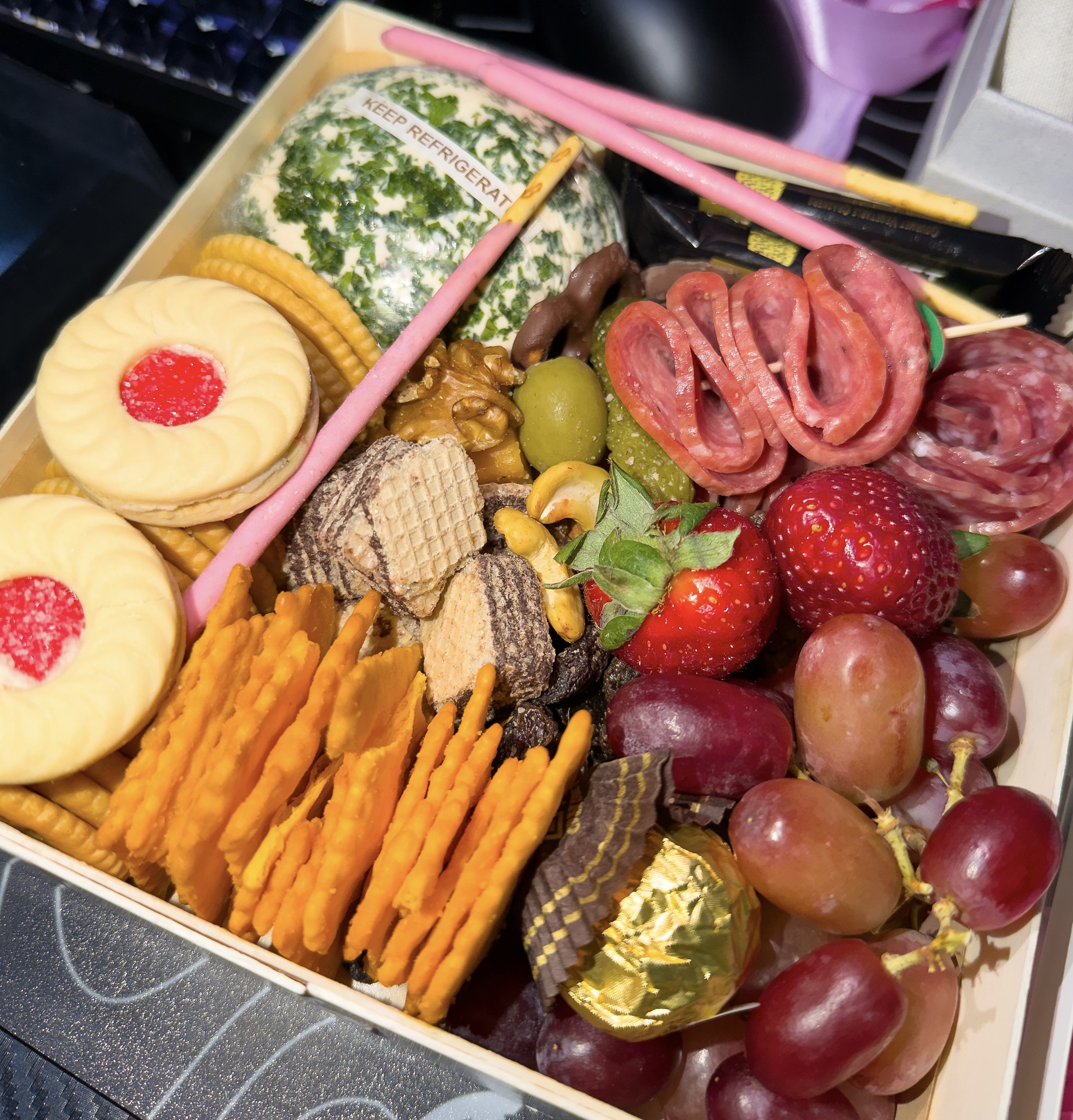 A square snack box filled with an assortment of sweet and savory treats. It includes folded salami, a herb-coated cheese ball, grapes, strawberries, various crackers, jam-filled cookies, wafer bites, pink Pocky sticks, and a gold-wrapped chocolate truffle.