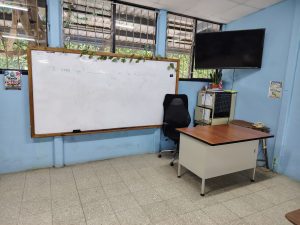 Whiteboard with and english grammar exercise written on it, next to a tracer desk and a tv monitor