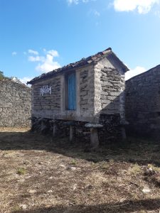 Rustic stone building with a blue door, tiled roof, and logs stored underneath.
