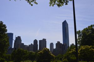 View from a park with lush green trees framing a skyline of modern and historic high-rise buildings.  A tall glass residential towers stand prominently against a clear blue sky