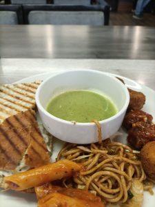 
A white plate filled with various foods. On one side, there is a slice of grilled bread with a ridged pattern, revealing a spread inside. Next to it, a small white bowl contains a vibrant green sauce.
