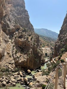 
A narrow canyon stretches between steep, rocky cliffs under a bright blue sky. The rocks are rugged and craggy, with patches of greenery scattered across their surfaces. 