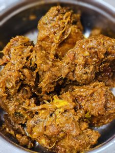 A close-up view of chicken pieces cooked in a thick, richly spiced gravy with onions and herbs. The warm brown and orange tones highlight the dish's texture and give the photograph a rich, homestyle feel. Captured in Koolimadu, Kozhikode, Kerala.