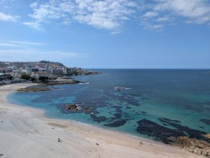 Panoramic view of Orzán and Riazor beaches in A Coruña with turquoise ocean waters, sandy coastline, and the city skyline under a blue sky.