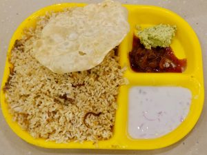 A plate of Malabar Chicken Biryani is served with papad, vegetable chutney, pickle, and creamy raita on a bright yellow plate. The contrasting colors and simple arrangement create a cheerful food photograph. Captured in Palazhi, Kozhikode, Kerala.