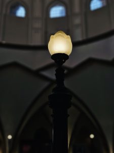 A glowing lotus-shaped lamp stands in a dark interior, creating a beautiful contrast between the warm light and surrounding shadows. The blurred architectural details and repeated lights add depth and atmosphere. Captured at Chhatrapati Shivaji Maharaj Vastu Sangrahalaya, Mumbai, Maharashtra.
