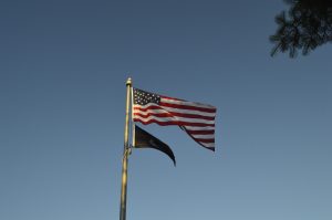 United States flag waving on a tall flagpole against a clear blue sky. A black flag hangs below it on the same pole, and the upper corner of a tree branch extends into the top-right of the image
