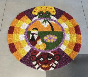 A colorful circular floral rangoli featuring a traditional figure surrounded by vibrant petal patterns.