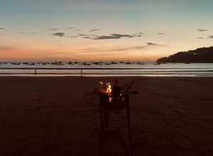 A beach scene at sunset, with a small fire burning in a grill at the foreground. The sky is painted in shades of orange, pink, and blue as the sun sets over a calm ocean. Silhouetted against the vibrant horizon, several boats are anchored in the water, gently swaying with the waves. 