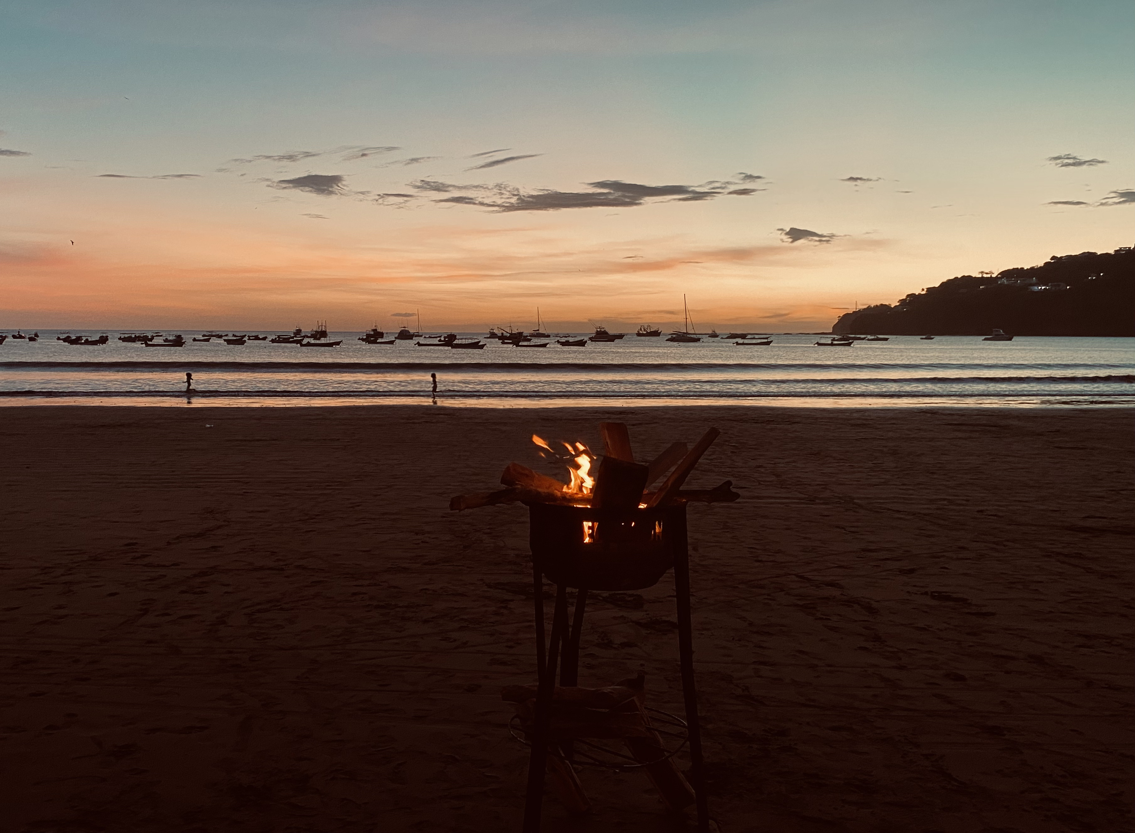 A beach scene at sunset, with a small fire burning in a grill at the foreground. The sky is painted in shades of orange, pink, and blue as the sun sets over a calm ocean. Silhouetted against the vibrant horizon, several boats are anchored in the water, gently swaying with the waves.