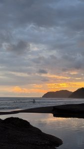 A surfer rides a wave at sunset on a beach with colorful skies.

