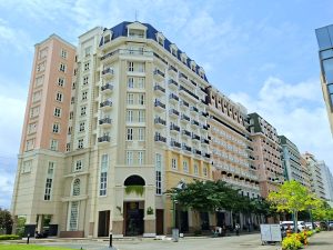 A row of multi-story European-inspired residential and commercial buildings featuring pastel facades, balconies, and a mansard roof under a bright sky.