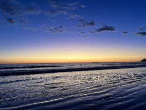 A serene beach scene at sunset, the sky transitions from deep blue at the top to warm shades of orange and yellow near the horizon. Wispy clouds scatter across the sky, reflecting the soft colors of twilight. The water is calm, with gentle waves rolling toward the shore, their surface shimmering like glass.