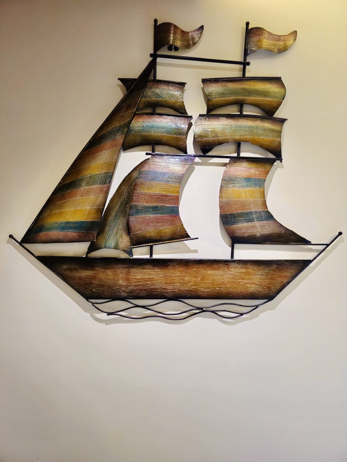 A decorative wall-mounted sailing ship features several curved golden-brown sails, a long wooden hull, and black metal supports. The warm colors and clean background make the detailed artwork stand out clearly. Captured in Chalappuram, Kozhikode, Kerala.