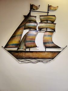 A decorative wall-mounted sailing ship features several curved golden-brown sails, a long wooden hull, and black metal supports. The warm colors and clean background make the detailed artwork stand out clearly. Captured in Chalappuram, Kozhikode, Kerala.