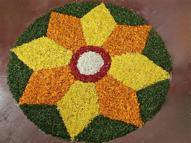 A colorful circular floral design (Onam Pookalam) made with fresh flower petals forms a large flower-like pattern. Bright yellow and orange petals surround a red and white center, while green petals form the outer border. Captured in Perumanna, Kozhikode, Kerala.