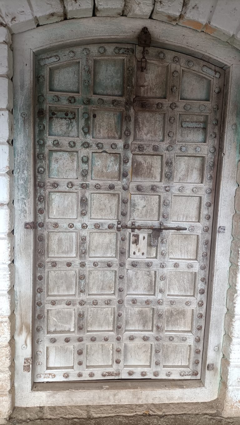 Old arched wooden door with faded blue and white paint, metal studs, and a rusted lock in a stone wall.