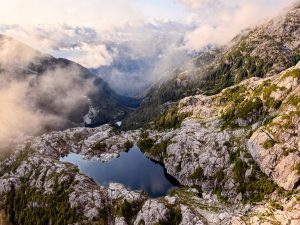 Aerial view of a dark alpine lake set in pale rocky terrain dotted with evergreens, with mist drifting over a deep glacier-carved valley and layers of mountain ridges beyond, lit by warm low-angle light.
