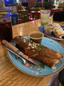 A crispy fried burrito topped with chopped onion and cilantro on a light blue plate, served with a small container of green salsa, alongside iced drinks on a wooden table in a colorful restaurant.