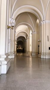 Interior corridor with high arches, pale-colored walls, and decorative columns along both sides. Wall-mounted lamps illuminate the tiled floor, leading toward a brightly lit area at the end of the hallway