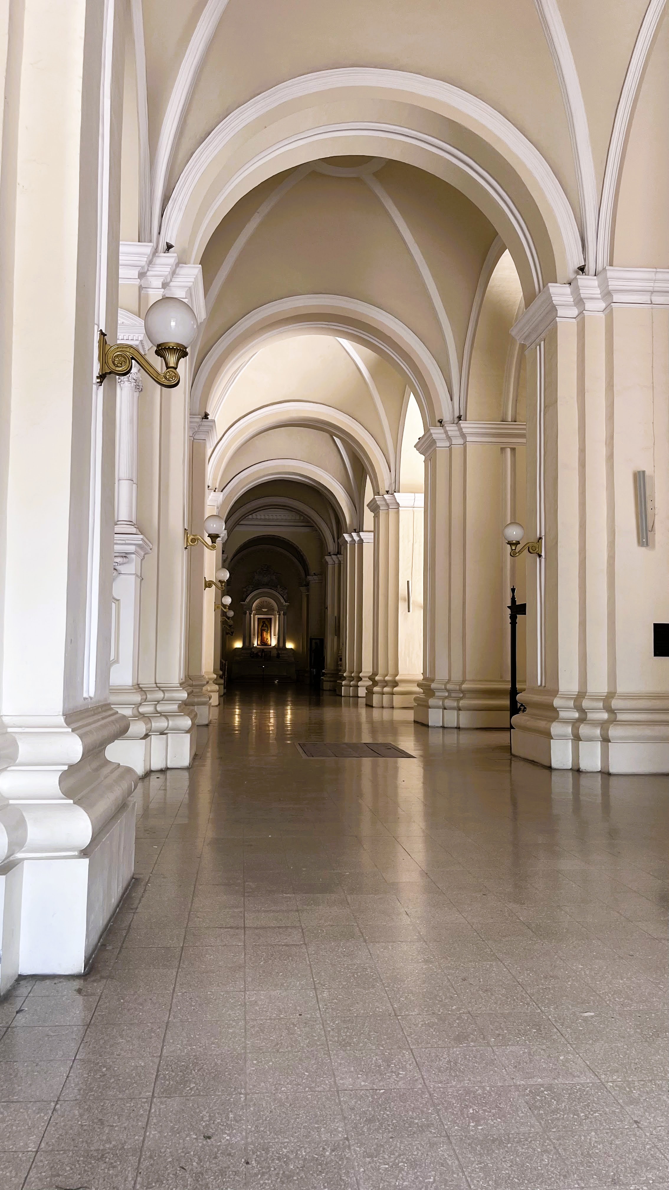 Interior corridor with high arches, pale-colored walls, and decorative columns along both sides. Wall-mounted lamps illuminate the tiled floor, leading toward a brightly lit area at the end of the hallway