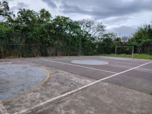 Outdoor concrete sports court at a highscool, surrounded by green vegetation and a trees under a cloudy sky
