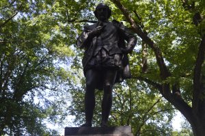  A statue of William Shakespeare standing on a stone pedestal beneath a canopy of leafy trees