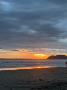 A serene beach scene at sunset, where the horizon is adorned with vibrant colors of orange and yellow as the sun dips into the ocean. The sky above is filled with soft, rolling clouds in varying shades of gray and blue. Gentle waves lapping at the shore create a soothing sound, while patches of wet sand reflect the warm hues of the sunset.