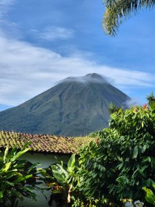 Lush green volcano partly covered by clouds, with tropical plants and a traditional house in the foreground under a clear blue sky.