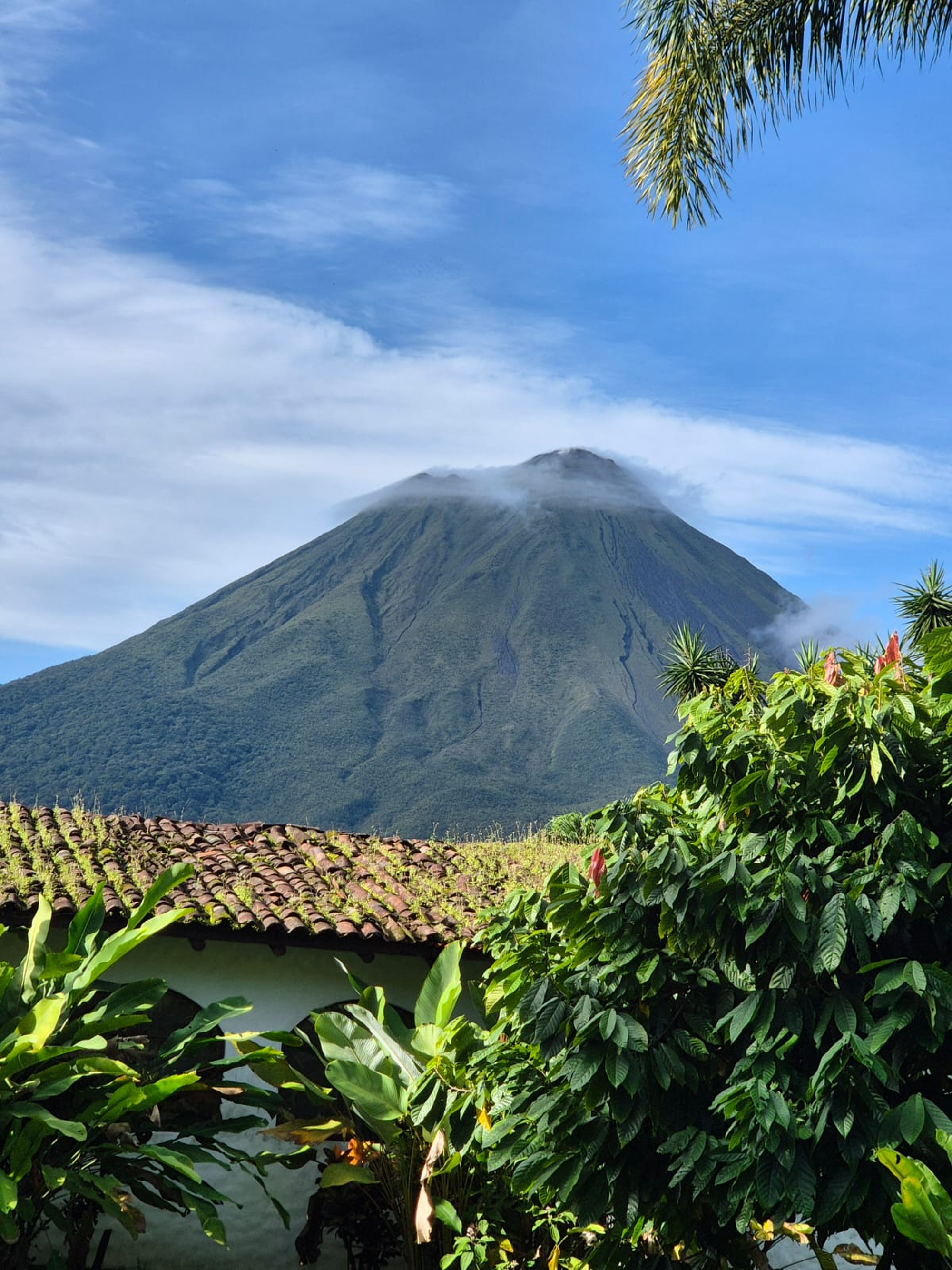 Lush green volcano partly covered by clouds, with tropical plants and a traditional house in the foreground under a clear blue sky.