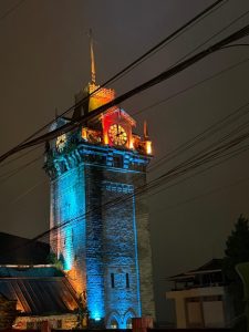 A tall clock tower illuminated at night, showcasing a mix of blue and orange lights that highlight its architectural details.