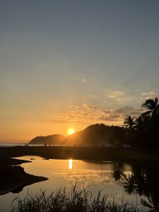 
Sunset over a calm beach with golden reflections on the water and a green hillside.