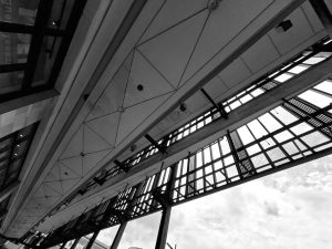 Low-angle view looking up at the geometric ceiling structure and dark metal framing of a modern building canopy against a cloudy sky.