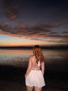 A woman with long, wavy red hair stands with her back to the camera, gazing at a beautiful sunset over the ocean. 