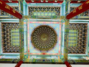 A colorful view of the ornate temple ceiling shows a large circular centerpiece surrounded by repeating patterns, dragons, flowers, and decorative beams. The strong symmetry and vivid colors highlight the beauty of Chinese temple architecture. Captured at Thean Hou Temple, Kuala Lumpur, Malaysia.