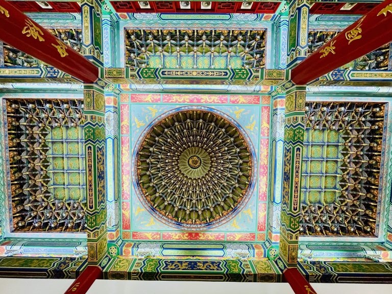 A colorful view of the ornate temple ceiling shows a large circular centerpiece surrounded by repeating patterns, dragons, flowers, and decorative beams. The strong symmetry and vivid colors highlight the beauty of Chinese temple architecture. Captured at Thean Hou Temple, Kuala Lumpur, Malaysia.