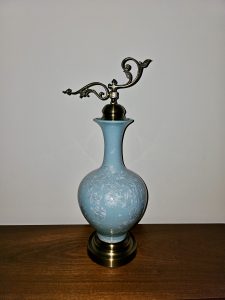 A light blue decorative vase with floral patterns and an ornate metal stopper stands on a wooden surface. Its rounded shape, delicate decoration, and curved metal finial create an elegant composition. Captured in Chalappuram, Kozhikode, Kerala.