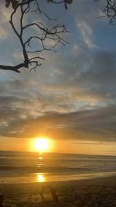A beach scene at sunset, where the vibrant orange and yellow hues of the setting sun reflect off the waves of the ocean. The sky is a mix of soft clouds with shades of blue and gray, creating a tranquil atmosphere.