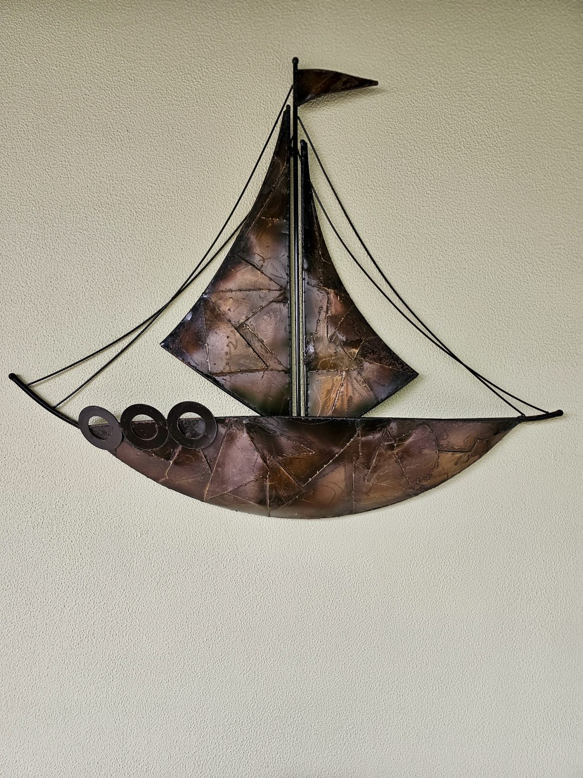 A handcrafted metal sailing boat is mounted against a textured light-colored wall. The sculpture has curved sails, a detailed hull, and thin rigging lines that create a simple nautical composition. Captured in Chalappuram, Kozhikode, Kerala.