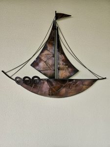 A handcrafted metal sailing boat is mounted against a textured light-colored wall. The sculpture has curved sails, a detailed hull, and thin rigging lines that create a simple nautical composition. Captured in Chalappuram, Kozhikode, Kerala.