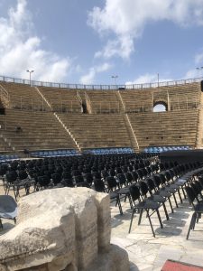 An ancient amphitheater is viewed from a low angle, showcasing a vast arrangement of empty black chairs arranged in rows on a stone floor. The seating is tiered, rising steeply toward the back, with a series of stairs leading up to the higher levels.