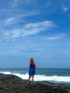
A woman with red hair standing on beach rocks by the ocean under a blue sky.