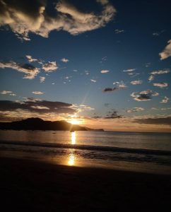 Sunset over a calm beach with golden light reflecting on the water and mountains in the distance.