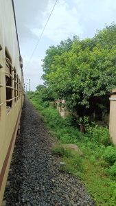 A beige and maroon train beside a railway track, with dense green foliage and an old building visible under a cloudy sky.