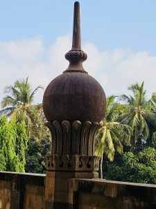 A large stone architectural ornament rises against a clear blue sky, with coconut palms and greenery in the background. Its rounded form, carved base, and tall finial create a strong vertical composition. Captured at Chhatrapati Shivaji Maharaj Vastu Sangrahalaya, Mumbai, Maharashtra.
