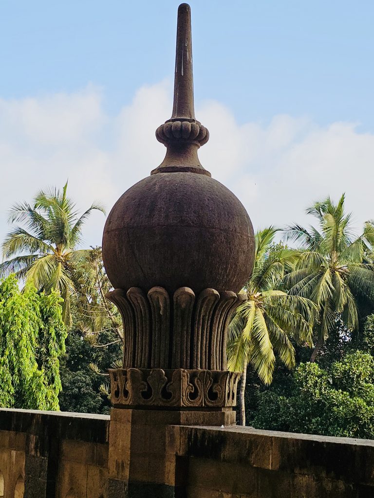 A large stone architectural ornament rises against a clear blue sky, with coconut palms and greenery in the background. Its rounded form, carved base, and tall finial create a strong vertical composition. Captured at Chhatrapati Shivaji Maharaj Vastu Sangrahalaya, Mumbai, Maharashtra.